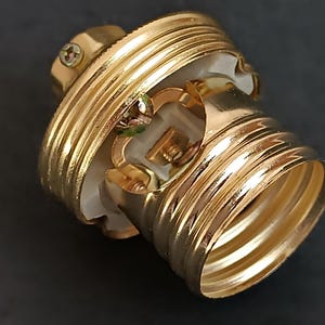 May include: Gold-toned light bulb socket with a white ceramic interior. The socket has a threaded base and a screw-on cap.