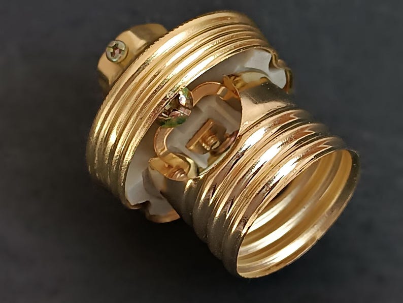 May include: Gold-toned light bulb socket with a white ceramic interior. The socket has a threaded base and a screw-on cap.