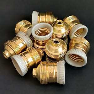 May include: A group of ten gold-coloured light bulb sockets with white ceramic bases. The sockets are arranged in a pile on a black surface.