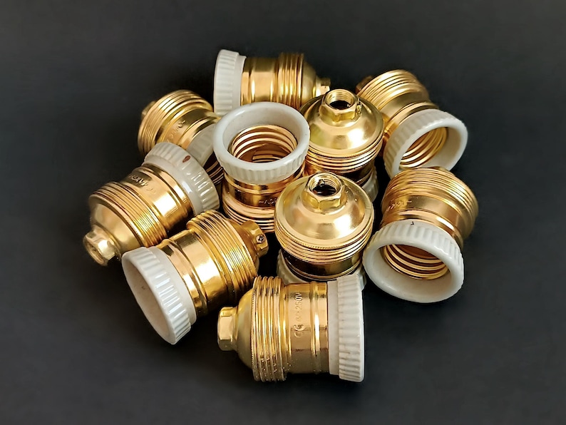 May include: A group of ten gold-coloured light bulb sockets with white ceramic bases. The sockets are arranged in a pile on a black surface.