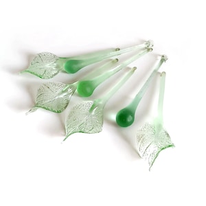 May include: A collection of delicate, light green glass leaf-shaped ornaments. Each ornament features a detailed leaf design and a rounded, teardrop-shaped stem. The glass has a translucent quality, and the overall aesthetic is elegant and nature-inspired.