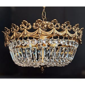 Peut inclure: Un lustre doré avec un design en forme de couronne. Le luminaire présente des cristaux transparents et un cadre orné. Le lustre est suspendu à une chaîne et est destiné à l'éclairage intérieur.