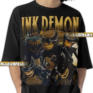 Puede incluir: Camiseta negra con texto dorado "INK DEMON" y un gráfico de varios personajes de Ink Demon. El gráfico presenta personajes con cuernos y dientes afilados. La camiseta tiene el texto "RusticGiftsArt".