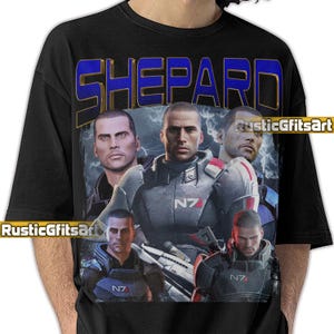 May include: Black t-shirt with a graphic of a man in a futuristic military uniform. The word "SHEPARD" is printed in blue and gold above the image. The image also includes the text "N7" in white.