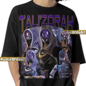 Puede incluir: Camiseta negra con un gráfico del personaje Tali'Zorah de la serie de videojuegos Mass Effect. El diseño incluye el nombre del personaje en letras moradas y doradas, con múltiples imágenes del personaje en un entorno futurista.