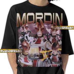 May include: Black t-shirt featuring the word "MORDEN" in gold lettering above a graphic of several alien figures in white and red uniforms. The image has a purple and white lightning background.