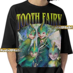 Może przedstawiać: Czarny t-shirt z grafiką czterech wróżek zębów z zieloną i niebieską skórą, skrzydłami i włosami. Tekst "TOOTH FAIRY" jest wygięty u góry w kolorze złotym i niebieskim. Koszulka ma okrągły dekolt.