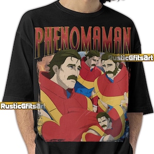 May include: Black t-shirt featuring a graphic of a man in a red and yellow outfit with the word "PHENOMANAN" in gold lettering. The graphic has a vintage, comic book style.