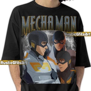 May include: Black t-shirt featuring a graphic of three figures in futuristic armor with the text "MECHA MAN" in a distressed font. The figures are in shades of blue, gray, and yellow, with a lightning background. The brand name "RusticGfitsArt" is also visible.