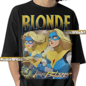 May include: Black t-shirt with a graphic of a blonde woman in a blue mask and yellow top. The word "BLONDE" is printed in gold above the image, and "Blazer" is printed in gold below.