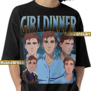 May include: Black t-shirt featuring the text "GIRL DINNER" in a blue and gold gradient. The shirt has a graphic of a man in a blue shirt, repeated multiple times. The graphic has a blue lightning background.