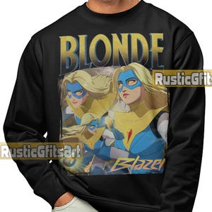 Puede incluir: Sudadera negra con un gráfico de una superheroína rubia con máscara azul y cuello amarillo. La palabra "BLONDE" está impresa en oro sobre la imagen. La palabra "Blazer" está impresa en azul debajo.