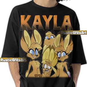 May include: Black t-shirt featuring a cartoon graphic of four anthropomorphic characters with large ears and blue eyes. The word "KAYLA" is printed in orange above the graphic. The text "RusticGfitsArt" is printed below the graphic.