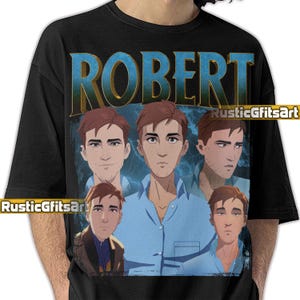 Peut inclure: T-shirt noir avec un motif graphique portant le nom "ROBERT" en lettres bleues et dorées. Le motif comprend plusieurs illustrations d'une personne aux cheveux bruns et portant une chemise bleue. Le texte "RusticGfitsArt" est également visible.