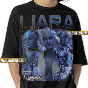 May include: Black t-shirt with a graphic of a blue-skinned character, the name "LIARA" in gold lettering, and multiple images of the character. The design includes the name "T'Soni" in a script font and the text "RusticGfitsArt".