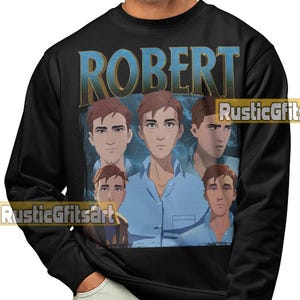 May include: Black sweatshirt featuring a graphic of a person with the name "ROBERT" in blue letters. The graphic includes multiple images of the person in a blue shirt. The sweatshirt has long sleeves and a crew neck.