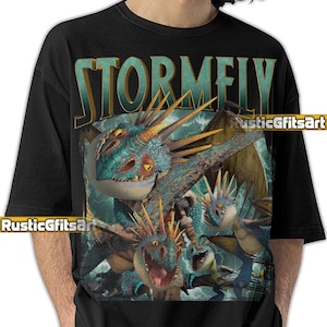 May include: Black t-shirt featuring a detailed graphic of dragons with the word "STORMFY" in teal. The dragon illustration includes teal, blue, and yellow colors. The shirt has a crew neck and short sleeves.