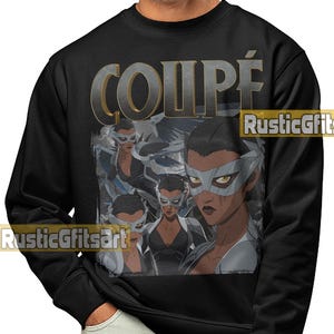 May include: Black sweatshirt featuring a graphic of a woman wearing a white mask and a black top. The word "COUPÉ" is printed in a metallic gold and gray font above the graphic. The sweatshirt is a crew neck style.