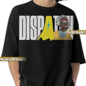 May include: Black crew neck t-shirt with the word "DISPATCH" in large white block letters. A cartoon illustration of a man wearing a headset and glasses is on the right. A yellow cape with a blue star is in the center.