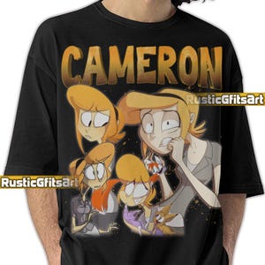 May include: Black t-shirt featuring the name "CAMERON" in gold lettering above a cartoon illustration of a woman with blonde hair in various poses. The design includes the text "RusticGfitsArt".