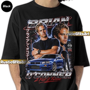 May include: A black t-shirt featuring a graphic print of Paul Walker as Brian O'Conner from 2 Fast 2 Furious. The design includes a blue Nissan GT-R, a tachometer, Japanese text, and the name Brian O'Conner in a metallic, chrome-style font.