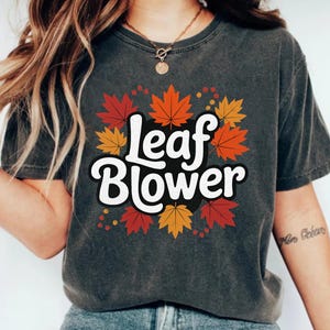 Peut inclure: T-shirt gris foncé avec l'inscription "Leaf Blower" en blanc, entourée de feuilles d'automne colorées. Les feuilles sont rouges, oranges et jaunes. Un t-shirt décontracté sur le thème de l'automne.
