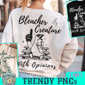 May include: A cream-colored sweatshirt with the text "Bleacher Creature With Opinions" and a graphic of a goose wearing sunglasses and holding a megaphone. The goose is sitting on a bench and wearing a foam finger that says "#1 FAN".