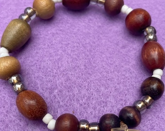 Puede incluir: Un brazalete de cuentas con cuentas de madera en tonos marrones y dorados, intercaladas con pequeñas cuentas plateadas y blancas. El brazalete está ensartado en un cordón elástico transparente, creando un aspecto natural.