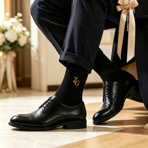 May include: A pair of black leather oxford shoes with a cap toe design and black laces, worn with black ribbed socks featuring the gold embroidered letters ZG. The shoes have a polished finish, a low heel, and a structured silhouette.