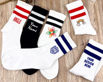 Puede incluir: Cinco pares de calcetines blancos de caña alta con rayas de colores y diseños diferentes. Un par tiene un arcoíris, uno tiene una flor, uno tiene un logotipo, uno dice "Your Name" y uno dice "Angle".