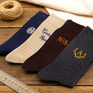 May include: Four ribbed socks in navy, beige, dark brown, and charcoal gray. The navy sock features a white hexagonal monogram, the beige sock has blue text reading Custom Name, the brown sock has orange text reading NIS, and the gray sock has a yellow antler emblem with the letter A.