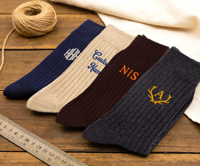 May include: Four ribbed socks in navy, beige, dark brown, and charcoal gray. The navy sock features a white hexagonal monogram, the beige sock has blue text reading Custom Name, the brown sock has orange text reading NIS, and the gray sock has a yellow antler emblem with the letter A.