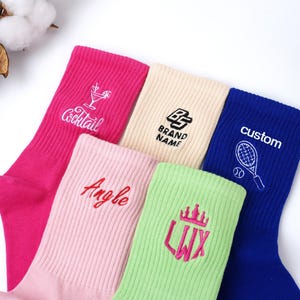 May include: A collection of colorful, ribbed crew socks. The socks are in shades of pink, cream, green, and blue. Each sock features embroidered designs or text, including a cocktail, a brand logo, the word "Angle", a crown, and the word "custom" with a tennis racket.