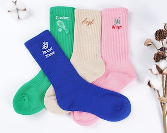 Puede incluir: Cuatro pares de calcetines coloridos con diseños bordados. Los calcetines son verdes, beige, rosas y azules. Los diseños incluyen una raqueta de tenis, la palabra "Angle", la palabra "Wifey" y un logotipo con las palabras "Brand Name".