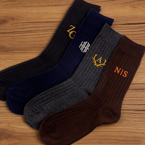 May include: Four pairs of ribbed socks in black, navy, gray, and brown, each embroidered with initials in gold or orange thread. The socks are arranged on a wooden surface.