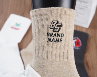 May include: Beige cotton socks with a black embroidered logo that says "BRAND NAME" and a stylized "BS" logo.