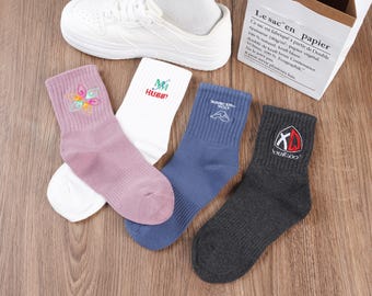 Puede incluir: Cuatro pares de calcetines con diferentes diseños bordados. Los calcetines son de varios colores, incluyendo rosa, blanco, azul y gris. Los diseños incluyen una flor, las palabras "Mi Hubby", una cordillera y las palabras "Your Logo".