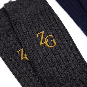 May include: A pair of dark charcoal gray ribbed socks featuring the embroidered gold letters ZG on the side. The fabric has a visible knit texture, and the socks are positioned next to a portion of a dark navy blue ribbed sock in the upper corner.