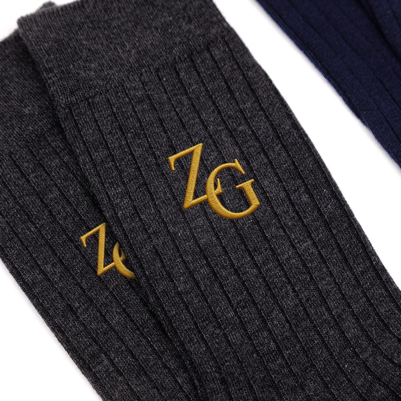 May include: A pair of dark charcoal gray ribbed socks featuring the embroidered gold letters ZG on the side. The fabric has a visible knit texture, and the socks are positioned next to a portion of a dark navy blue ribbed sock in the upper corner.