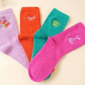 May include: Four pairs of colourful socks: lavender with a floral design, orange with the word "Angle", teal with a heart and "I love you" text, and pink with a cocktail glass design. The socks are made of a soft, fuzzy material.