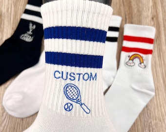 May include: Three pairs of white athletic socks with colorful stripes. One pair has a blue stripe and a tennis racket and ball design with the word "CUSTOM" embroidered on the sock. Another pair has a red stripe and a rainbow with clouds embroidered on the sock. The third pair has a black stripe.