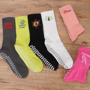 Könnte beinhalten: Sechs Paar bunte rutschfeste Socken mit verschiedenen Designs und Texten. Die Socken sind grau, gelb, schwarz, weiß, rosa und rosa mit einem Tennisracket-Motiv. Der Text auf den Socken lautet "Your Name", "Brand Name", "Pilates" und "Custom".