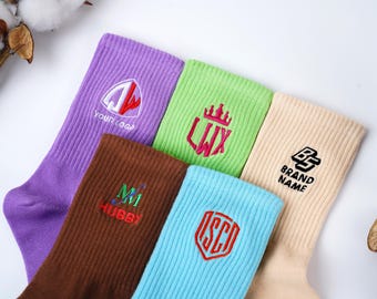 Puede incluir: Seis pares de calcetines coloridos con logotipos y texto bordados. Los calcetines son en tonos de morado, verde, marrón, azul claro y beige. Cada calcetín presenta un logotipo o texto único, incluyendo "YOUR LOGO", "LWX", "MM HUBBY", "ISCO" y "BRAND NAME".
