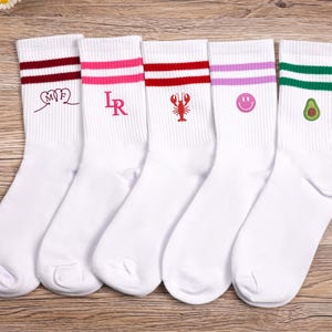 Könnte beinhalten: Fünf Paar weiße Sportsocken mit farbigen Streifen und gestickten Designs. Die Socken haben bordeauxrote, rosa, rote, lavendelfarbene und grüne Streifen. Designs sind Initialen, ein Hummer, ein Smiley und eine Avocado.
