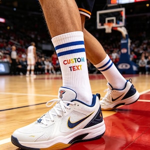 Puede incluir: Calcetines deportivos blancos con rayas azules y el texto "CUSTOM TEXT" en colores arcoíris. Los calcetines se usan con zapatillas deportivas blancas y azul marino con un logotipo dorado, en una cancha de baloncesto.