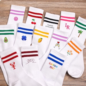 Könnte beinhalten: Eine Sammlung weißer Crew-Socken mit gerippten Bündchen mit zwei farbigen Streifen und verschiedenen gestickten Symbolen, darunter ein Smiley, Kirschen, Avocado, Kleeblatt, Mango, Palme, Hummer, Blumen sowie der Text Custom Embroidery oder Custom Text.