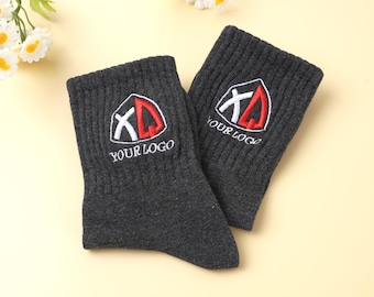 May include: A pair of charcoal gray socks with a ribbed texture. Each sock features an embroidered logo in white, red, and black, with the text "YOUR LOGO" below the logo. The socks are displayed on a light yellow surface.