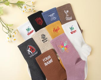 Puede incluir: Una colección de calcetines deportivos en varios colores, cada uno bordado con diseños y textos únicos. Los calcetines presentan textos como "HUBBY", "WIFEY", "YOUR LOGO" y "YOUR NAME", junto con otros gráficos. Los calcetines están dispuestos sobre una superficie amarillo claro.