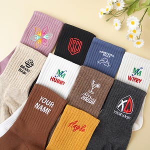 May include: A collection of colorful socks with embroidered designs and text. The socks come in various colors, including black, white, brown, and blue. Some socks feature the text "YOUR NAME", "WIFEY", and "HUBBY". Other socks have logos and floral designs.