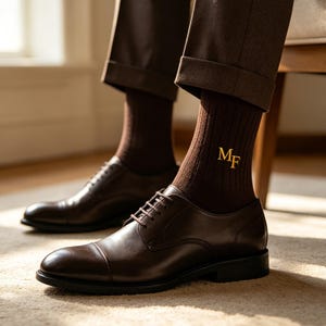 May include: A pair of dark brown leather cap-toe oxford shoes paired with dark brown ribbed socks. The socks feature the embroidered gold-colored letters MF on the outer side. The shoes have a smooth finish, thin laces, and a low heel.